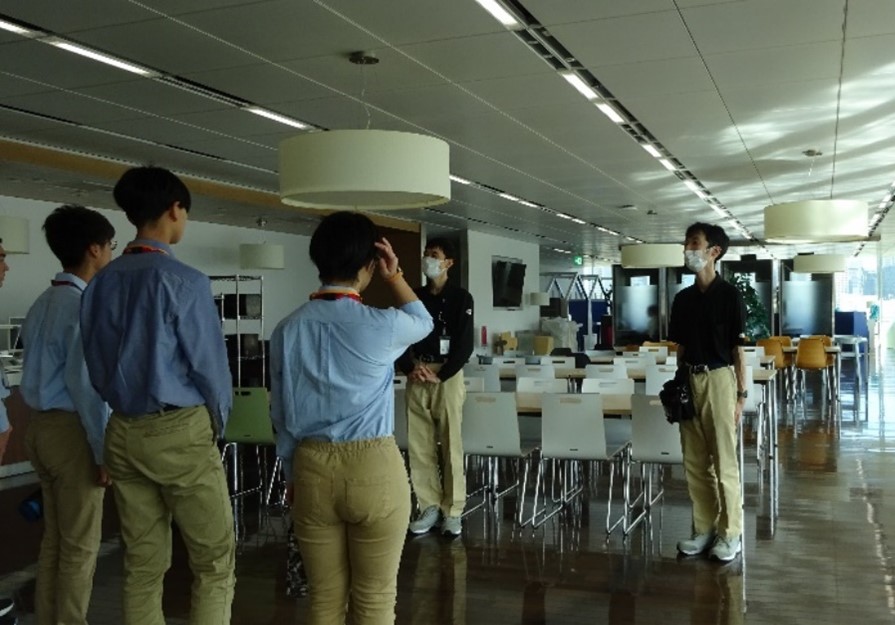 社員食堂のような広いスペースで、複数の人が立って話している。白い椅子と長テーブルが整然と並び、天井に円形照明がある。